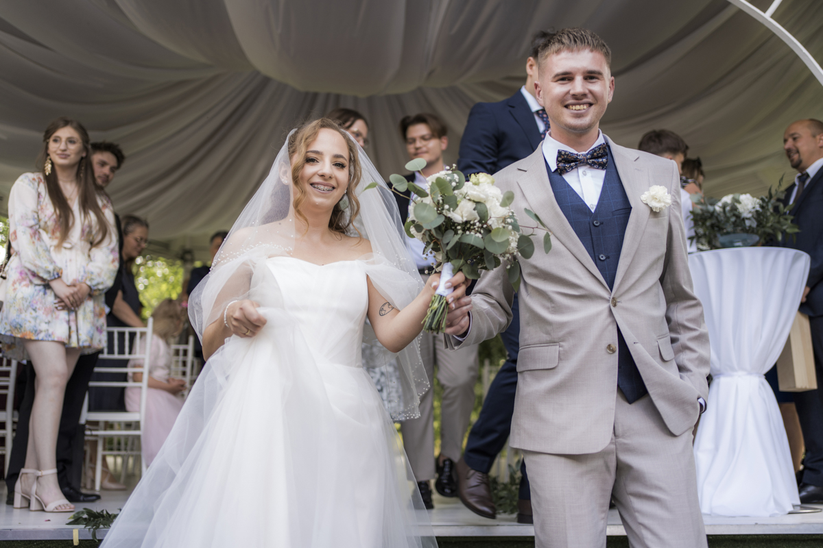 Para młoda uśmiechnięta opuszcza miejsce ceremonii w Zajeździe pod Kasztanami, w tle biała dekoracja i goście